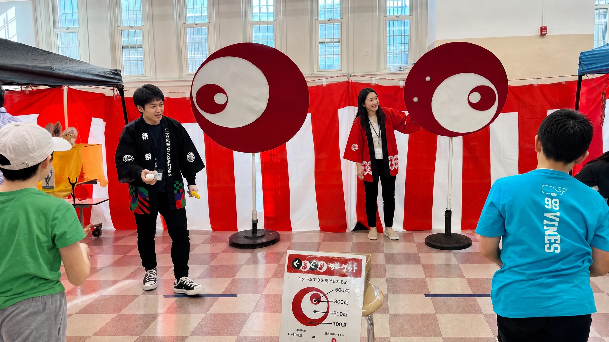 Children throwing balls at two spinning eye-shaped target discs in front of red-and-white bunting