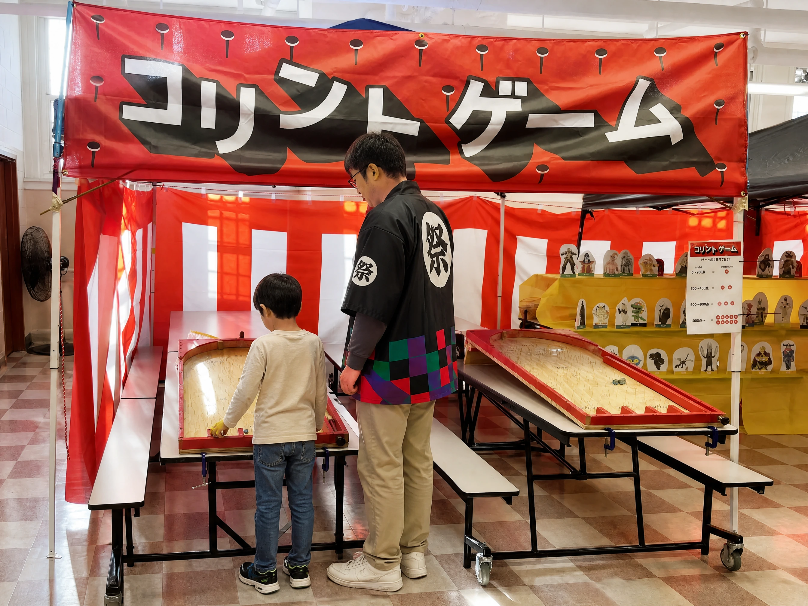 A single child standing on the floor, back to the camera, playing Korinto while an adult volunteer watches beside them