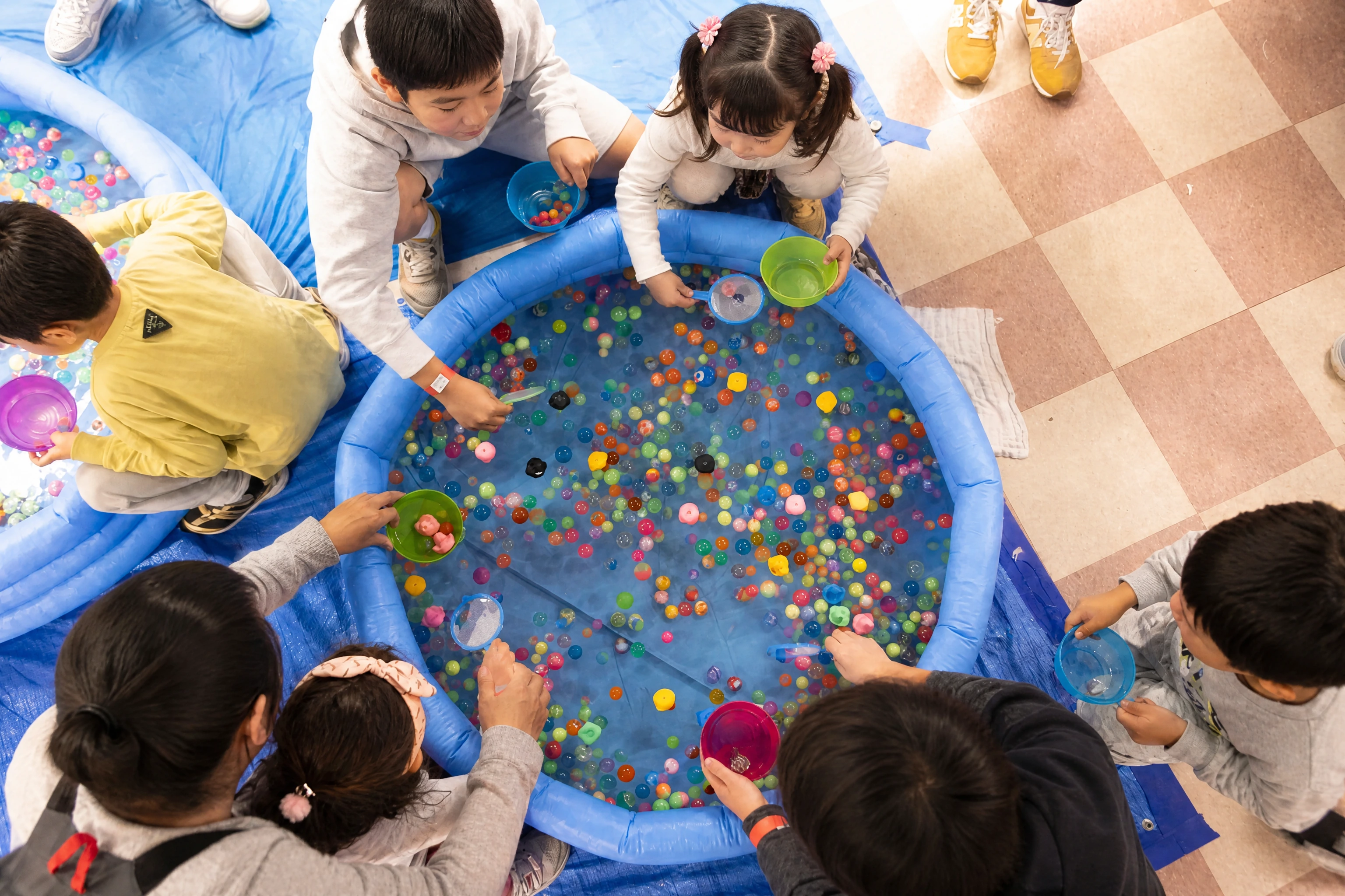 Overhead view of children around a paddling pool full of colourful super balls, scooping with paper nets and bowls